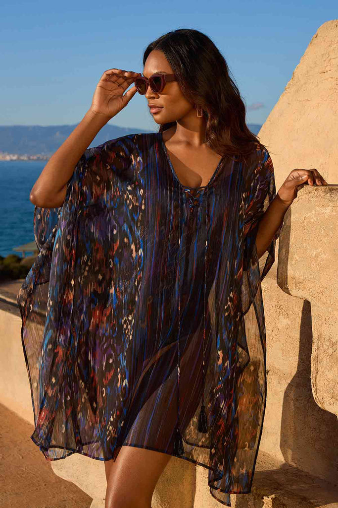 Woman facing left in sunglasses wearing a multicolored swim cover up in front of a beach background